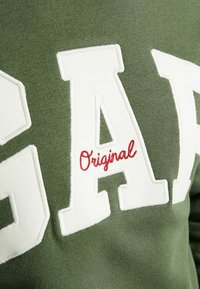 Zelená mikina s velkým bílým nápisem "GAP" a vyšitým "Original" v červené barvě. Měkká látka a ležérní design.