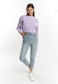 Fioletowy, krótki sweatshirt z okrągłym dekoltem i haftowanym napisem, w zestawieniu z jasnoniebieskimi dżinsami z wysokim stanem i białymi trampkami.