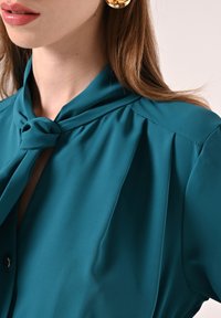 Blusa color turchese con colletto annodato, texture liscia e pieghe sulle spalle. Orecchini circolari dorati completano l'outfit.