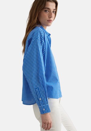 Mujer joven con una camisa de manga larga a cuadros azul y blanca y pantalones blancos, de pie de perfil con una expresión neutral.