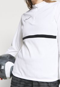 Weißes Nike Langarmoberteil mit einem halben Reißverschluss, das einen schwarzen horizontalen Streifen und eine glatte Textur aufweist, kombiniert mit einem schwarz-weißen Fußballs.