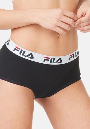 Gros plan d'une personne portant un short avec ceinture noire de la marque FILA, les mains ajustant la ceinture près du nombril sur un fond blanc.