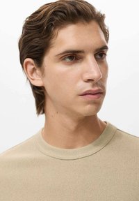 Tricou crem cu guler rotund, caracterizat printr-un material texturat, mâneci scurte și un guler ribbed, purtat de o persoană cu păr șaten deschis.
