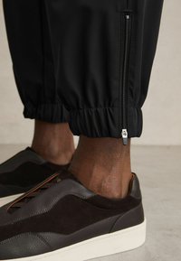Pantaloni jogger negri cu manșete elastice și detalii cu fermoar aproape de gleznă, asortați cu adidași maro închis, cu un design texturat și șireturi.