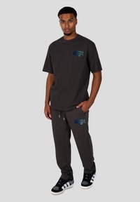 Dunkelgraues Baumwoll-T-Shirt mit kurzen Ärmeln, das mit blauer und grüner Grafikkunst gestaltet ist. Kombiniert mit passenden Jogginghosen und schwarzen Turnschuhen.