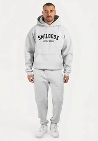 Felpa grigia con cappuccio, con logo nero stampato "SMILODOX ATHL. WEAR" sul davanti, abbinata a pantaloni della tuta grigi coordinati e scarpe da ginnastica bianche.