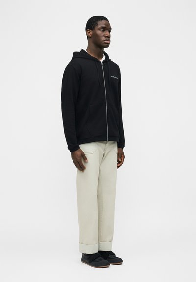 Άνδρας που στέκεται φορώντας μαύρο φούτερ με φερμουάρ, ανοιχτό μπεζ παντελόνι με φαρδιά μπατζάκια και μαύρα αθλητικά παπούτσια, με φόντο ένα απλό λευκό φόντο.