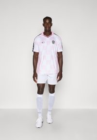 Lichtroze geruite voetbalshirt met zwarte accenten, witte shorts en witte sokken. Heeft een v-hals en een logo op de borst.
