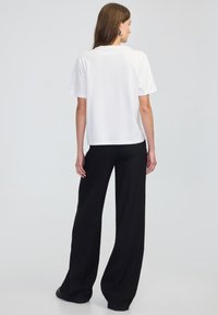 Vrouw met lang bruin haar, gekleed in een witte korte mouwtop, zwarte wijde broek en zwarte schoenen, staande tegen een lichte achtergrond.