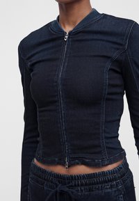 Personne portant une veste zippée bleu foncé ajustée et un pantalon à cordon de serrage assorti avec une taille élastique, visible du cou à la taille.