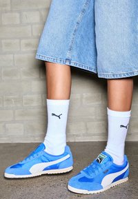 Ben klädda i ljusblå denimshorts, vita Puma-sockar och blå Puma-sneakers står på en grå betonggolv framför en tegelvägg.