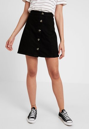 Jupe noire en denim coupe A avec taille haute, fermeture à boutons à l'avant et légère déchirure à l'ourlet. Associée à des baskets noires.