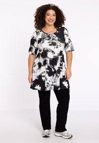 Zwart-wit gestreepte tuniek met korte mouwen en een uitlopende zoom, gedragen over zwarte broeken en sportschoenen. Glad fabric met bloemdesign.