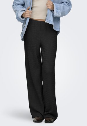 Person wearing high-waisted textured black wide-leg pants, cropped beige top, light blue denim jacket, and dark brown square-toe shoes.