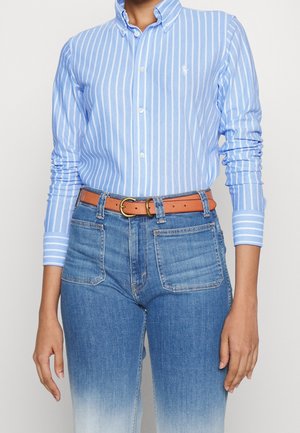 Persoon draagt een blauw-wit verticaal gestreept overhemd met knopen, ingestopt in blauwe high-waisted jeans met voorzakken en een bruine riem.