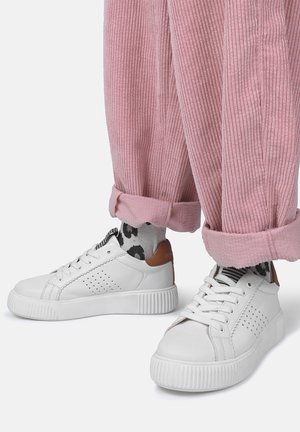 Hvide lave sneakers med perforerede sider og brun hælskinne, båret med sorte og hvide sokker med mønster og optrukne lyserøde manchesterbukser.