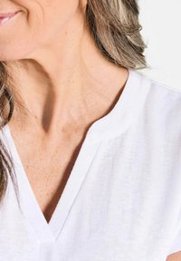Witte V-hals top van zachte stof, met een smalle kraag en minimale stikdetails. Close-up toont de huidskleur en haar dat zachtjes de nek omlijnt.