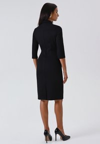 Donna in piedi, che indossa un abito nero aderente fino al ginocchio con maniche a tre quarti e scarpe col tacco alto nere, di spalle.