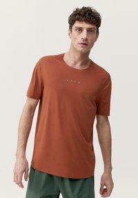 Hrdzavá krátkopytová tri čká vyrobené z ľahkého materiálu s okrúhlym výstrihom. Obsahuje jemný detail loga na prednej strane.