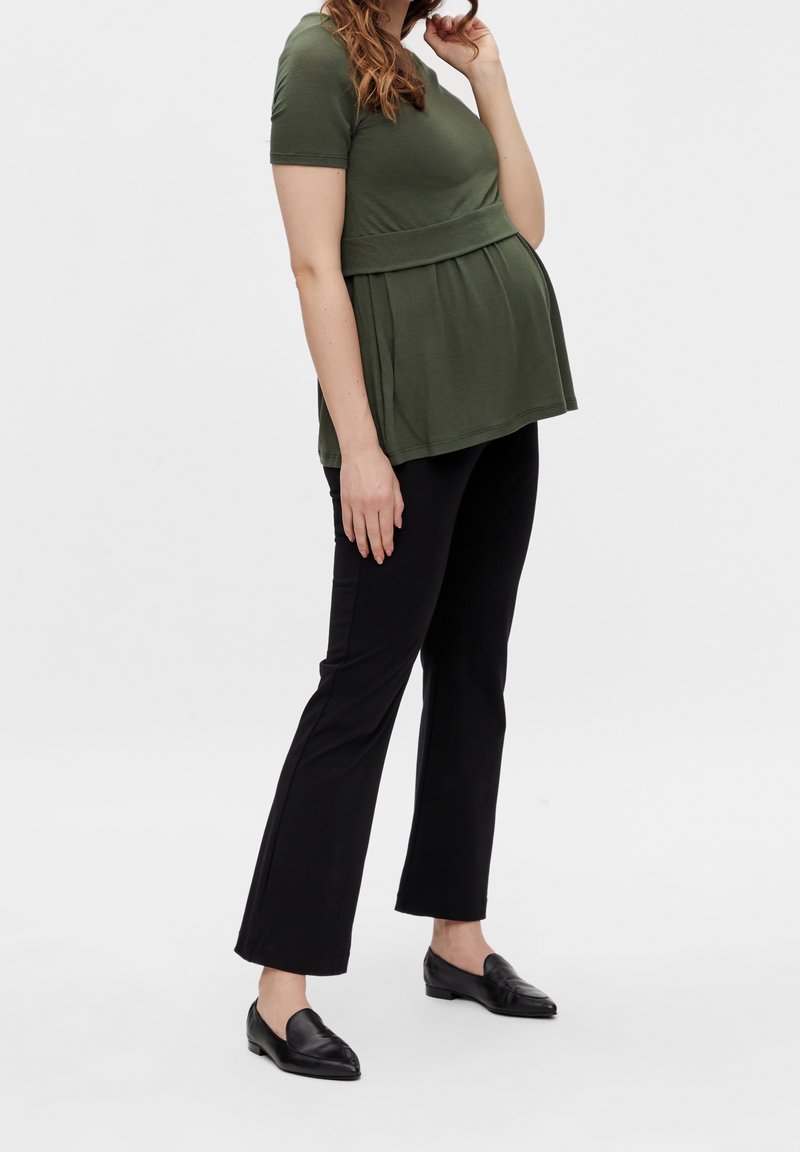 Pregnant woman wearing an olive green top, black pants, and black loafers standing against a plain white background.