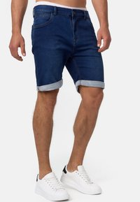 Un homme portant des shorts en denim roulés de couleur bleu foncé et des baskets blanches, se tenant devant un fond clair.