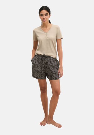 Mujer de pie descalza, vistiendo una camiseta beige de manga corta con cuello en V y pantalones cortos negros con estampado floral y cordón, con las manos en los bolsillos, fondo neutro.