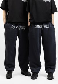 Pantalones de chándal azul marino en un corte relajado con un logo plateado de "Wasted" en la cinturilla, combinados con zapatillas deportivas negras.