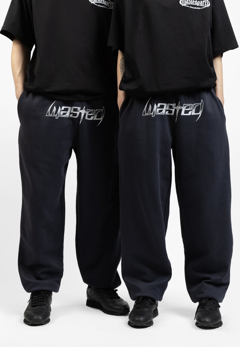 Pantalones de chándal azul marino en un corte relajado con un logo plateado de "Wasted" en la cinturilla, combinados con zapatillas deportivas negras.