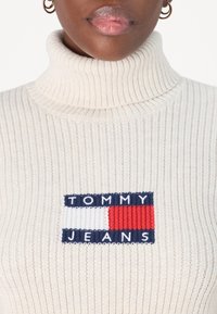 Maglione a collo alto lavorato a maglia di colore crema con texture a coste, dotato di una toppa logo rettangolare nei colori blu navy, rosso e bianco al centro del petto.