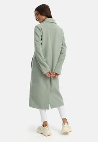 Lange mintgroene jas van fleece-stof met een brede kraag, zijsplitten en lange mouwen, gecombineerd met witte legging en chunky sneakers.