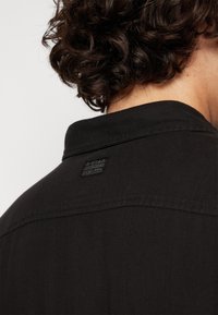 Μαύρο πουκάμισο, από στερεό ύφασμα, με γιακά και μια μικρή μαύρη ετικέτα λογότυπου στο πίσω μέρος. Δείχνει λεπτομέρειες ραφής και υφή.