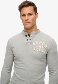 Hellgrauer Langarm-Sweatshirt mit einem gerippten Kragen, zwei braunen Knöpfen und beigem, gesticktem Text "SUPER DRY TOKYO" auf der Brust.