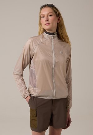 Joven con cabello largo y rubio, vestida con chaqueta beige con cremallera y pantalones cortos marrón oscuro con bolsillos laterales, de pie con una mano en el bolsillo.