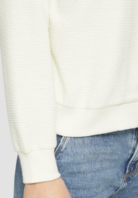 Pull blanc texturé avec un motif côtelé horizontal, doté de poignets et d'un ourlet côtelés, associé à un jean en denim bleu.