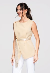 Femme portant un haut beige sans manches avec des détails à boutons et une ceinture nouée, associée à un pantalon blanc, souriant et regardant sur le côté.