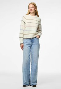 Mlada žena u kremastom puloveru s pastelnim prugama i svijetloplavim širokim trapericama stoji s jednom rukom u džepu.