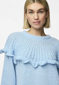 Svetlo modra pletena jopica z rebrastim vzorcem in volanastimi poudarki pri ovratniku. Mehka tekstura in sproščen kroj.