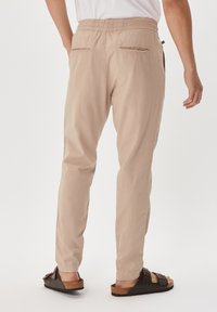 Beige, leichte Hose mit elastischem Bund, zwei Gesäßtaschen und taillierter Beinform. Getragen mit dunklen Sandalen.