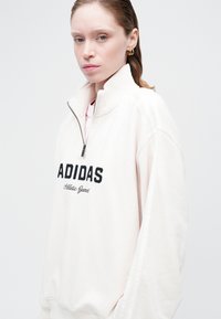 Felpa zip-up Adidas di un beige chiaro con logo nero, dotata di collo alto e accenti bianchi a righe sulle maniche. Texture morbida.