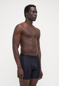 Fekete boxertartó, textúrált anyagból készült, márkázott elasztikus derékpánttal, testhezálló fazonban és a comb közepéig érő hosszal.