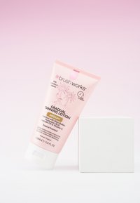 Lotion autobronzante Brush Works dans un tube rose, avec du texte blanc, à côté d'une boîte carrée blanche. Contient 150 ml, parfum tropical noté.