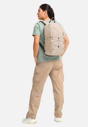 Frau trägt beigefarbene Cargohose, grünes kurzärmeliges Hemd, weiße Sneakers und einen beigen Rucksack mit elastischen Frontkordeln.