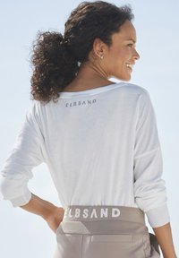 White long-sleeve top with a loose fit and wide neckline, featuring a subtle "ELBSAND" logo on the back. Light gray waistband with matching text.