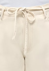 Μπεζ τζιν παντελόνι με θηλιές για ζώνη και δέσιμο υφασμάτινου κορδονιού στη μέση, που δείχνει λεπτομέρειες μπροστινής ραφής και μέρος ενός λευκού ριμπ πλεκτού τοπ.