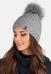Beanie grigio a maglia con texture a coste, dotato di un pom-pom grigio soffice in cima e di un piccolo etichetta in pelle marrone sul lato.