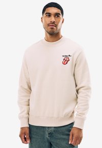 Ljusbeige sweatshirt med rund halsringning. Har en röd och svart Rolling Stones-logotyp på bröstet. Mjuk bomullsblandad tyg.