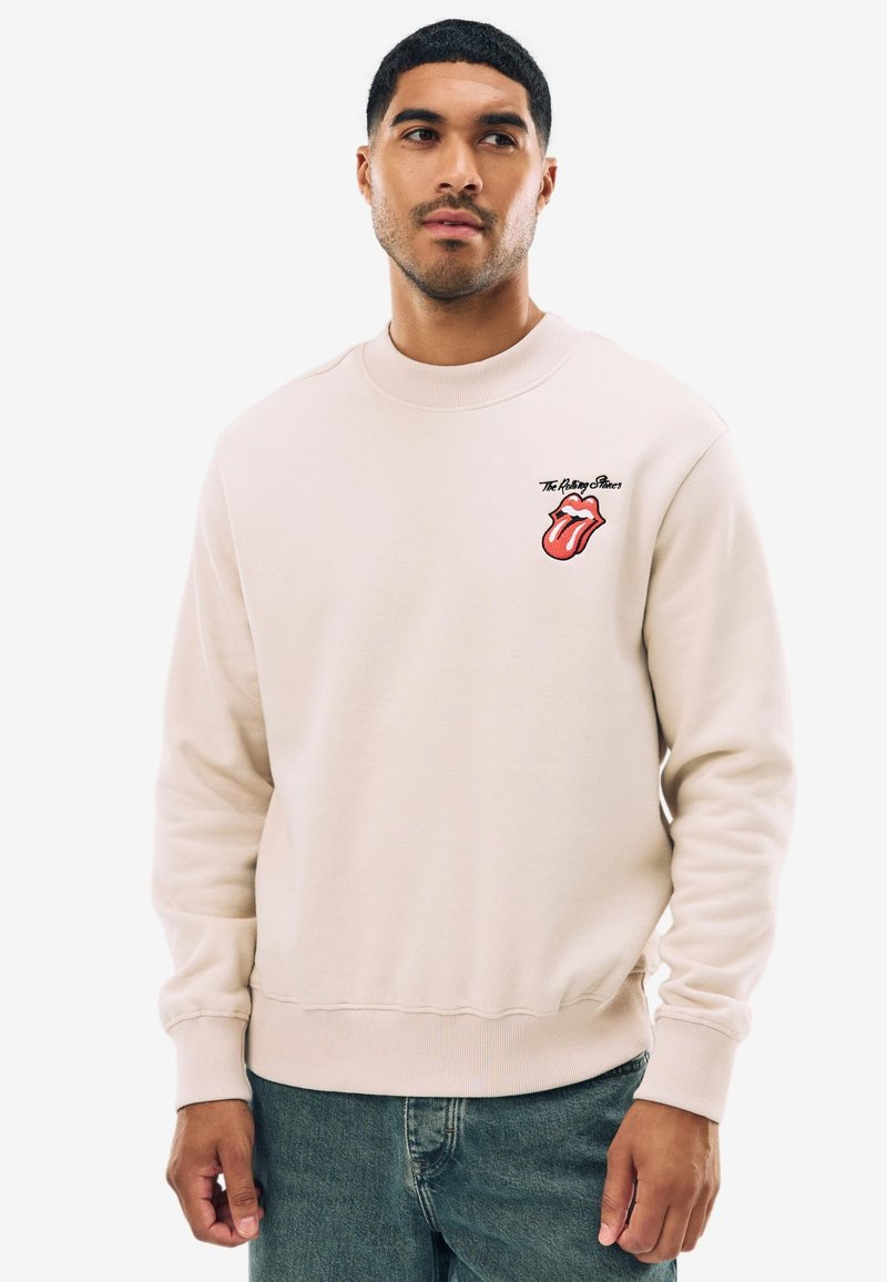 Ljusbeige sweatshirt med rund halsringning. Har en röd och svart Rolling Stones-logotyp på bröstet. Mjuk bomullsblandad tyg.