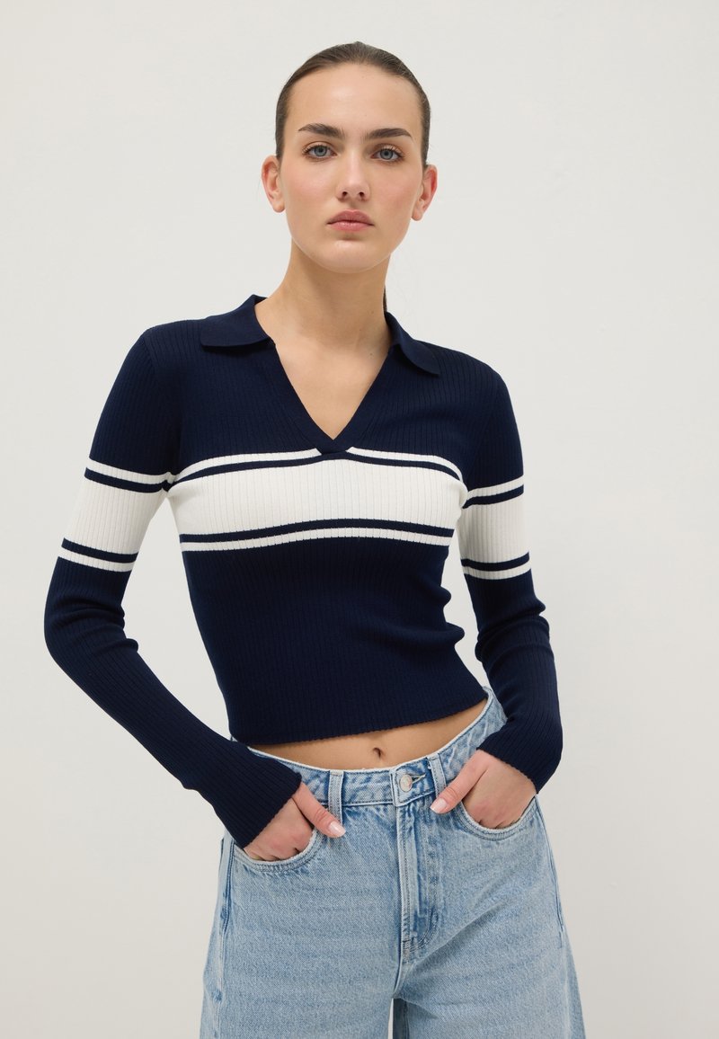 Marineblauwe geribbelde trui met witte en marineblauwe strepen, V-hals en korte kraag. Gecombineerd met lichtblauwe, hooggesneden jeans met een cropped pasvorm.