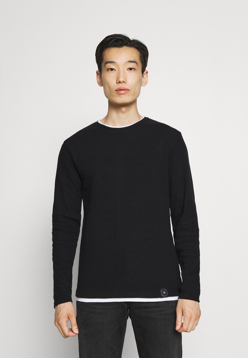 Chemise noire à manches longues avec un col rond, dotée d'un col intérieur blanc, d'un tissu texturé et d'un petit logo sur le côté inférieur.