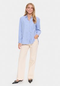 Camisa abotonada a rayas azul y blanca con detalles bordados, combinada con pantalones anchos color crema y zapatos de tacón negro de punta.